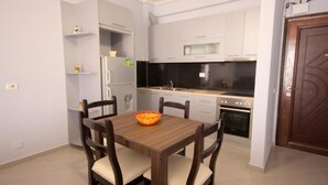 A compact kitchen with white cabinets, a wooden dining table, and a built-in oven. - Ideal Holiday Apartments (Sarande)