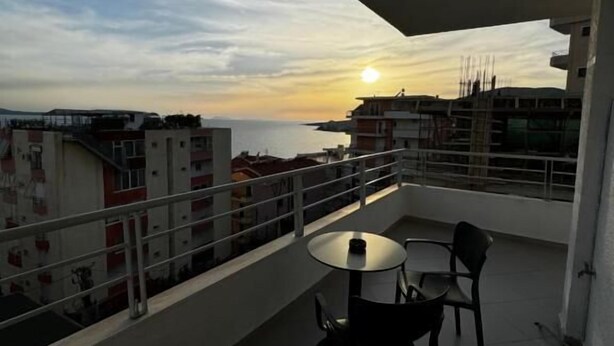 A balcony with a table and chairs, overlooking a cityscape and the sea during sunset. - Ideal Holiday Apartments (Sarande)