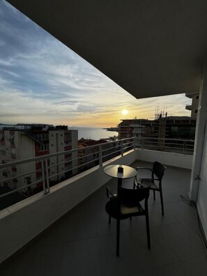 A balcony with a table and chairs, overlooking a cityscape and the sea during sunset. - Ideal Holiday Apartments (Sarande)