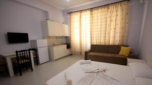A hotel room with a bed, a sofa, a dining table with chairs, a TV, a kitchenette, and curtains. - Ideal Holiday Apartments (Sarande)