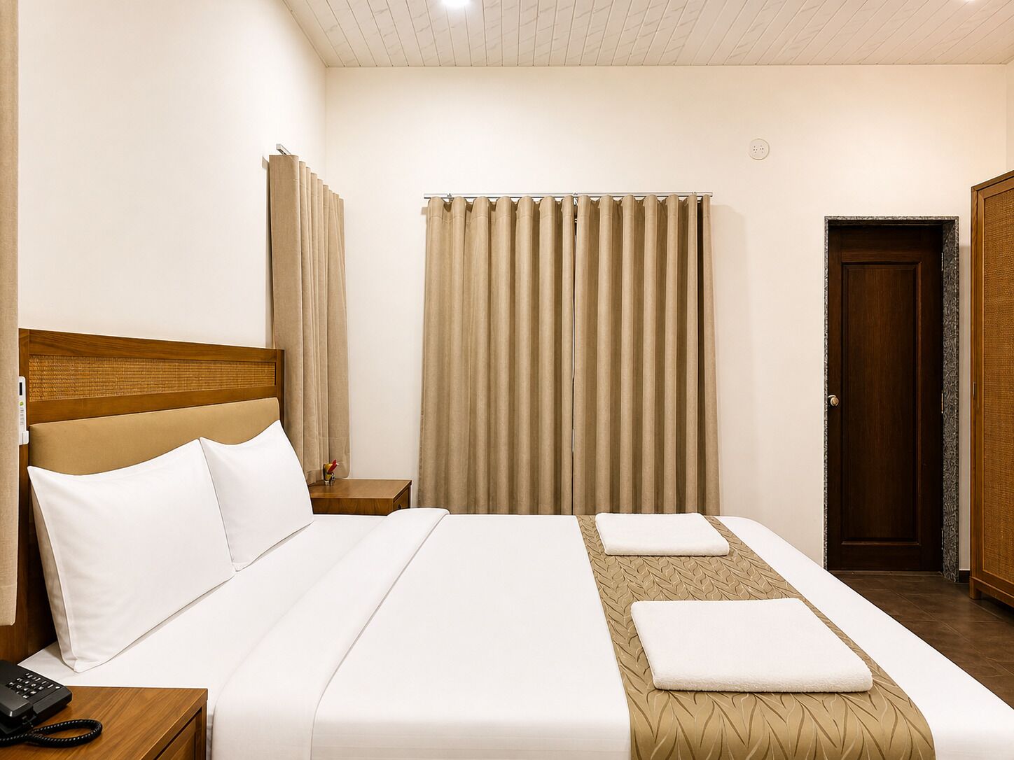 A hotel room with a bed, a bedside telephone, a wooden headboard, a window with curtains, and a door.