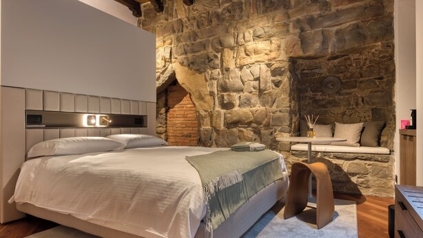 A bedroom with a stone wall, a bed with a headboard, a small table with a lamp, and a bench against the wall. - Le Torri Suites (Bergamo)