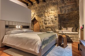 A bedroom with a stone wall, a bed with a headboard, a small table with a lamp, and a bench against the wall. - Le Torri Suites (Bergamo)