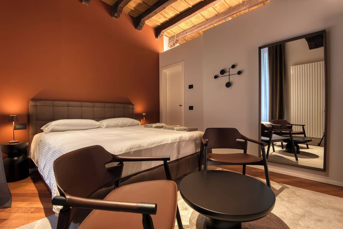 A modern hotel room with a large bed, two armchairs, a small round table, a nightstand, and a large mirror reflecting the room.