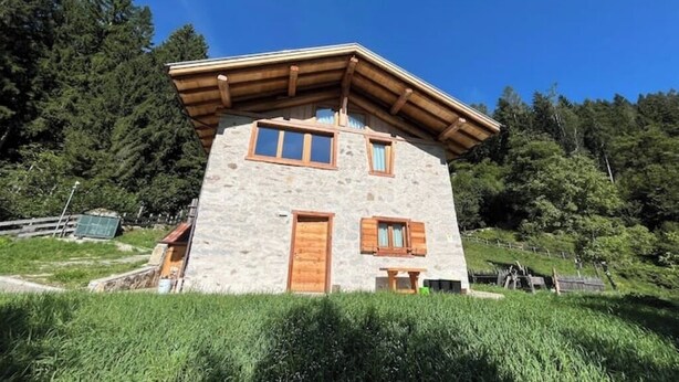 Una casa de piedra con techo inclinado y vigas de madera, rodeada de abundante follaje y árboles.