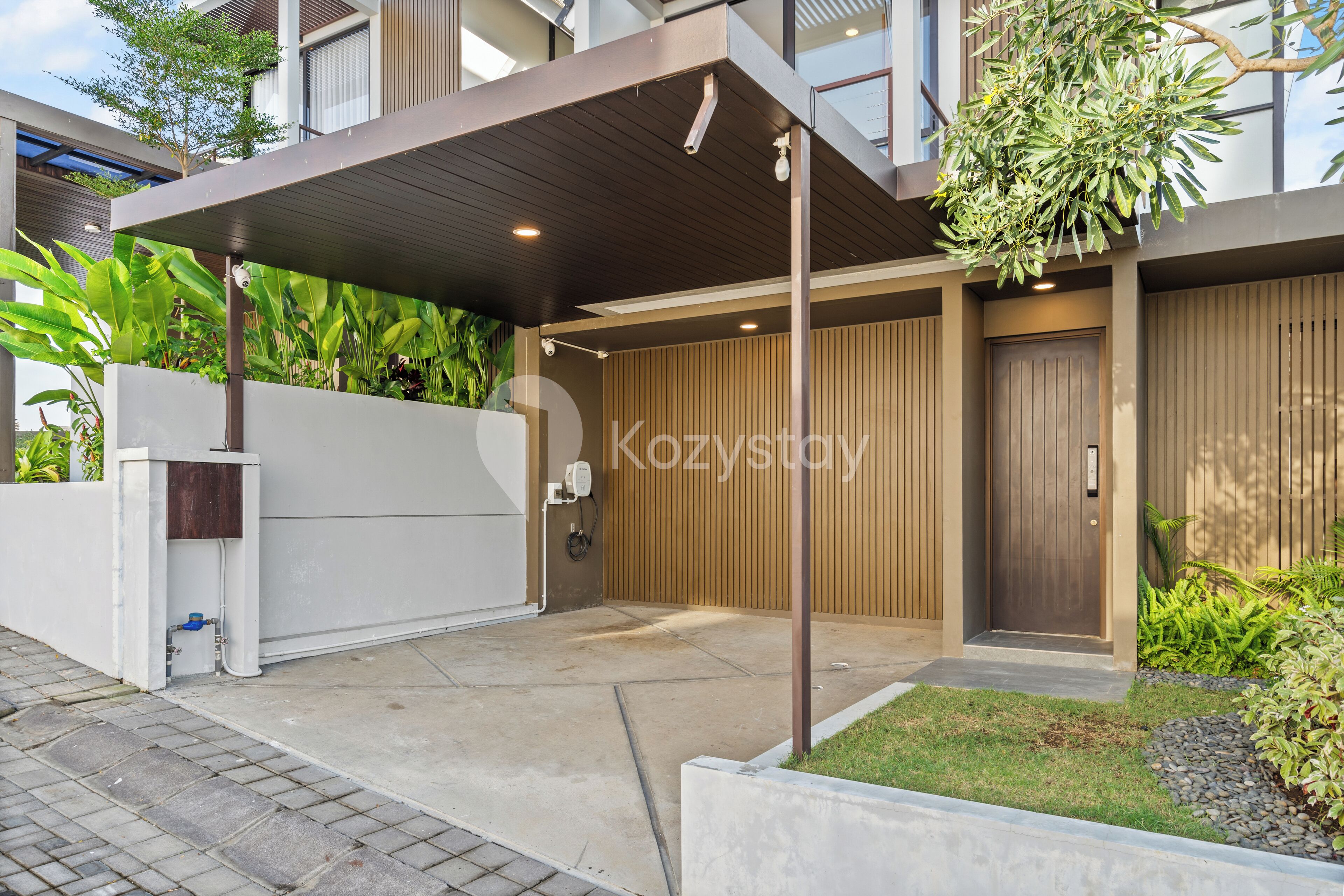 A modern building with a covered entrance, a wooden door, and a small garden area with plants.
