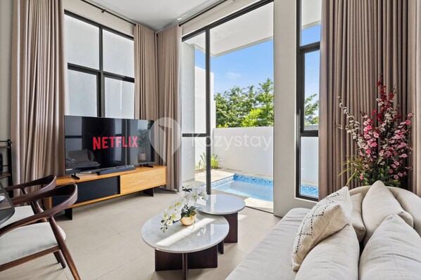 A modern living room with a flat-screen TV, a sofa, a coffee table, and a view of a swimming pool. - Dharma Villa by Kozystay - Jimbaran (Jimbaran)