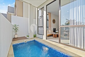A modern outdoor pool area with a glass door leading to a living room, a potted plant, and a view of the sky. - Dharma Villa by Kozystay - Jimbaran (Jimbaran)