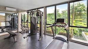 A modern gym with various exercise equipment, large windows offering a view of greenery, and a clean, spacious layout. - Dharma Villa by Kozystay - Jimbaran (Jimbaran)