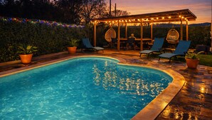 Une piscine agrémentée d’une pergola et de chaises, entourée de plantes en pot et illuminée de guirlandes lumineuses.