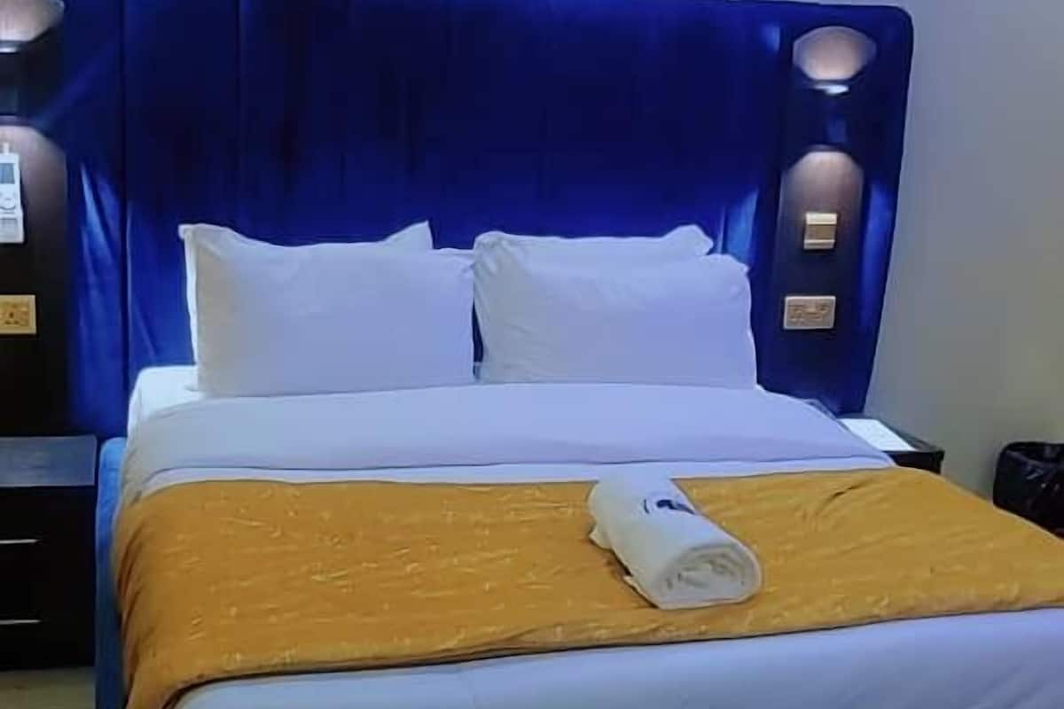 A hotel room with a large bed, white bedding, a blue headboard, and an air conditioning unit above the bed.
