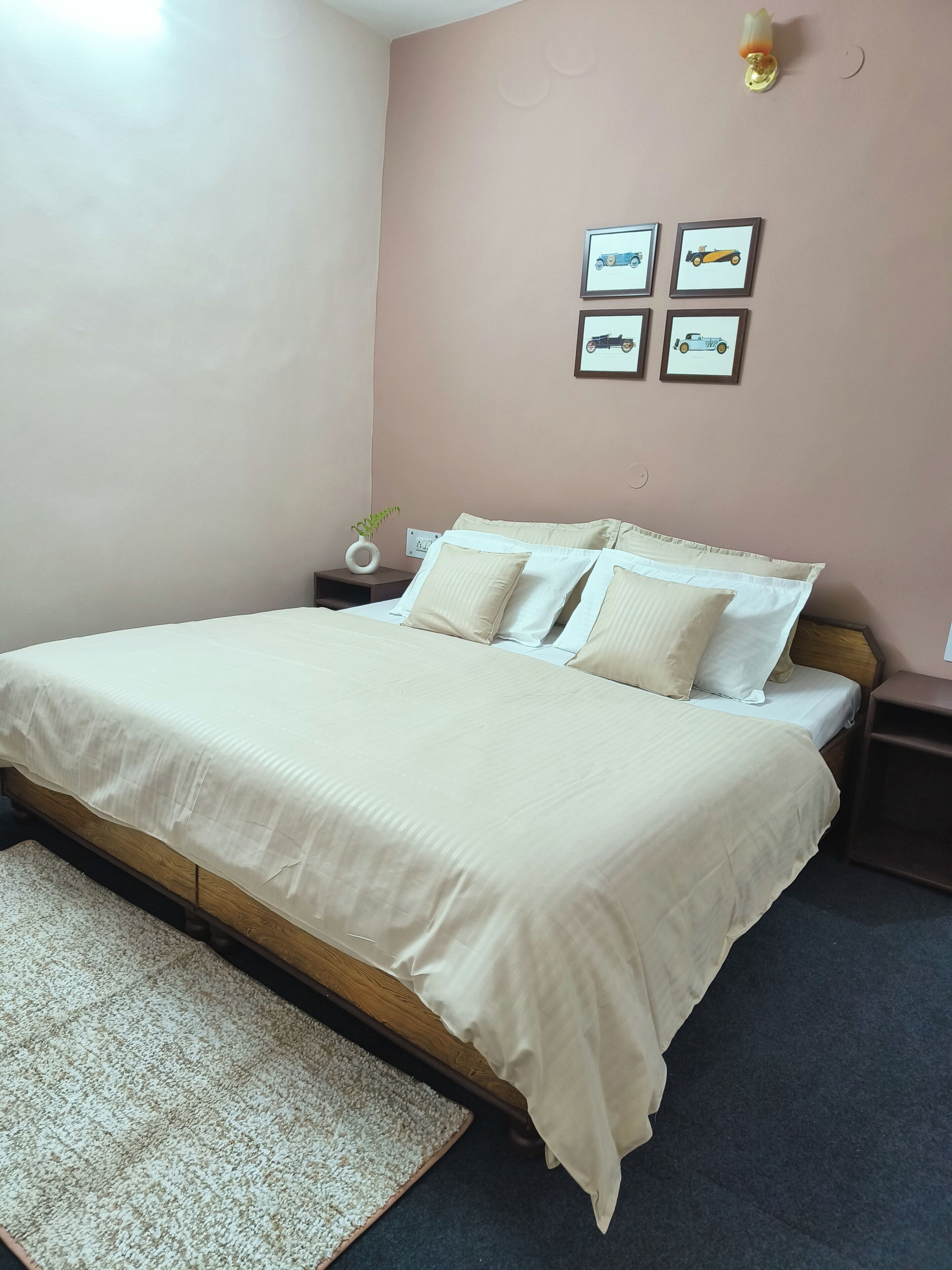 A neatly made bed with a white bedspread and pillows, a small bedside table with a lamp, and a wall with four framed pictures of vehicles.