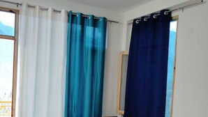 A room with a wooden floor, a ceiling fan, a table with chairs, and curtains in blue and white. - THE KAILASHA (Naggar)