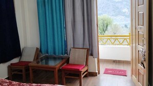 A room with a balcony, two wooden chairs, a glass table, and a view of mountains through the window. - THE KAILASHA (Naggar)
