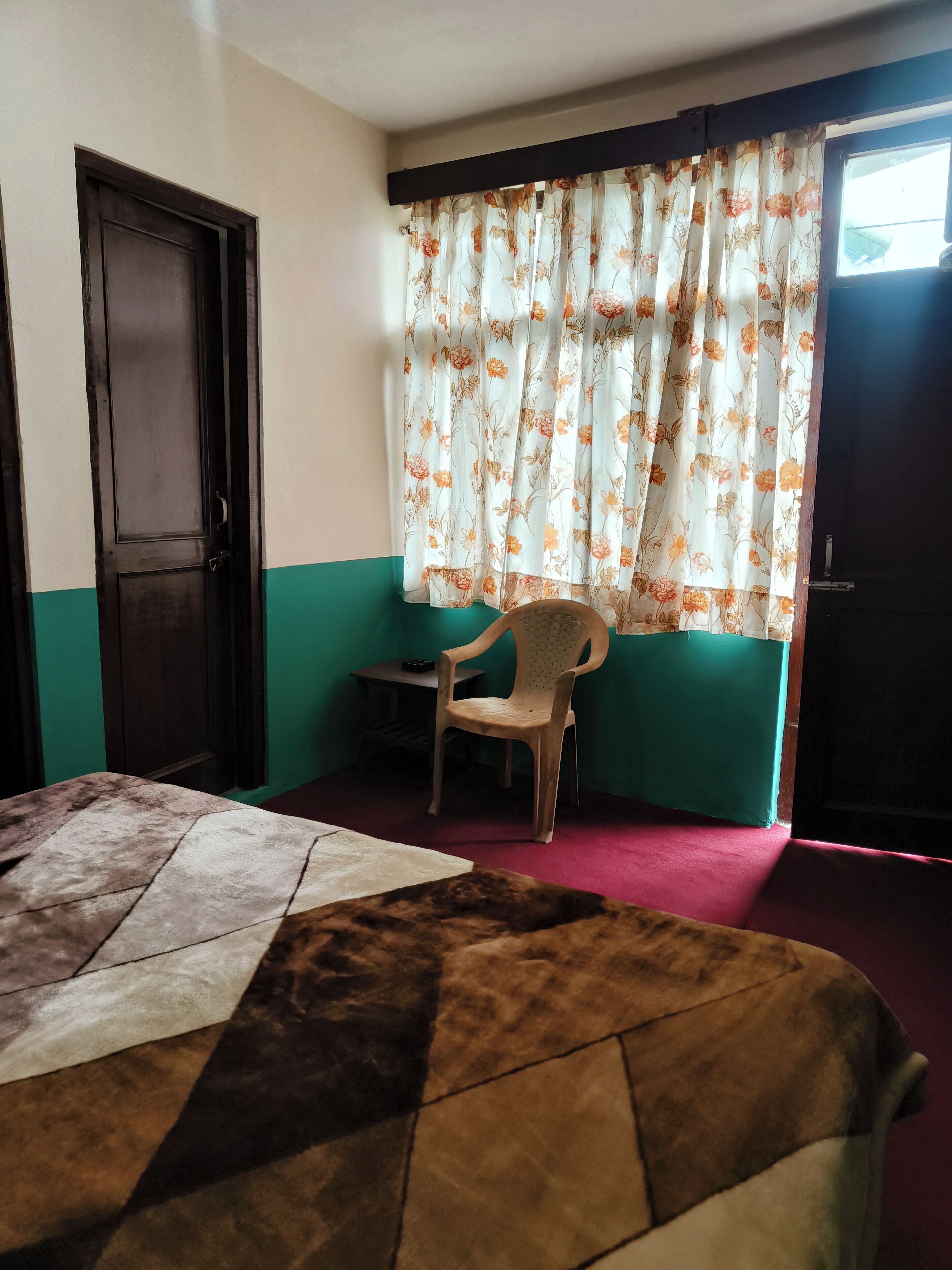 Una habitación con cama, una silla, una mesita y un ventanal con cortinas florales.