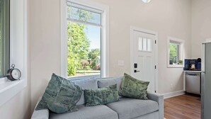 A modern living room with a grey sofa, green patterned cushions, a large window with blinds, a small kitchen area, and a door.