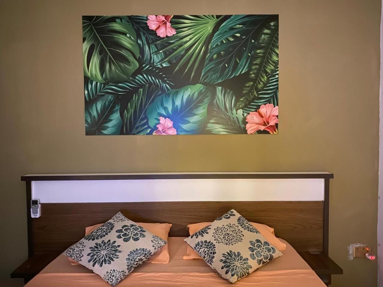Une chambre à coucher comprenant un lit, deux oreillers, une table de chevet et une œuvre d’art fixée au mur représentant des feuilles et des fleurs tropicales.