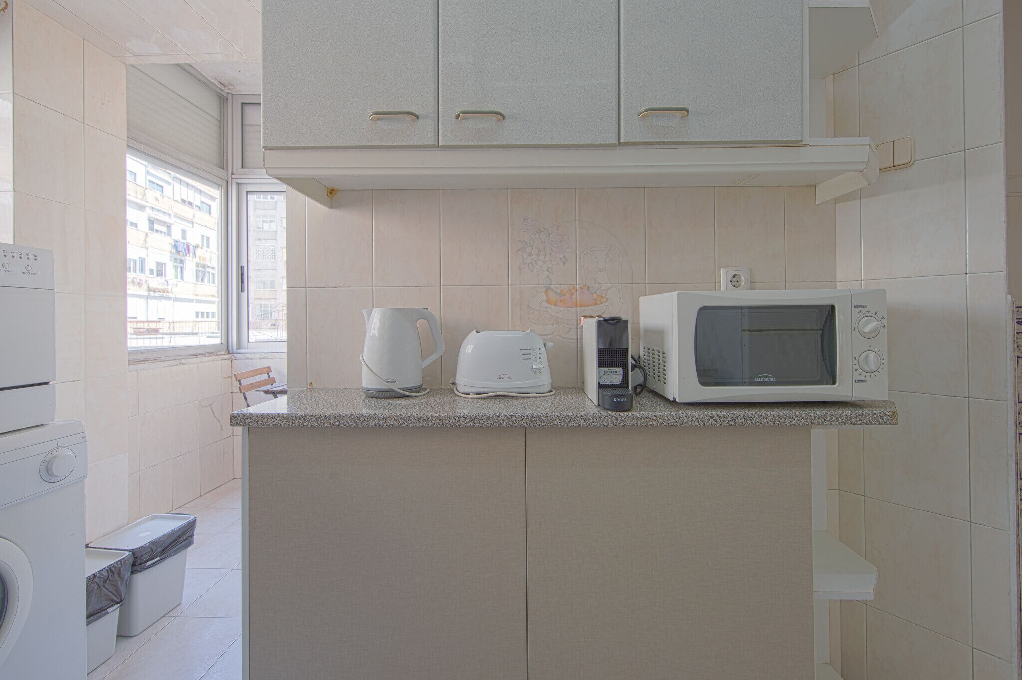 A kitchen with a microwave, kettle, and toaster on the counter, a washing machine, and a window with a view of buildings.
