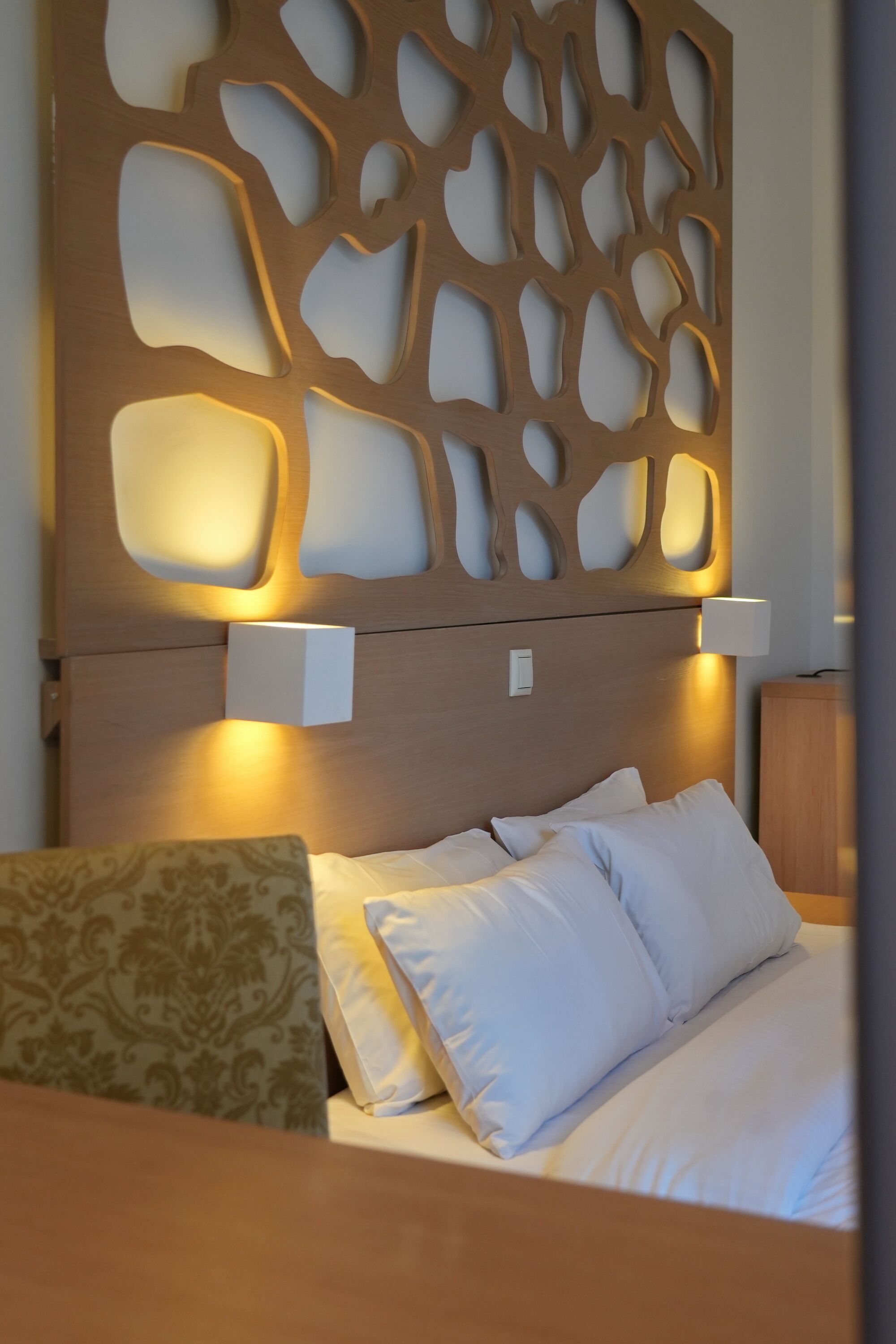 A hotel room with a wooden headboard, two bedside lamps, a bed with white linens, and a patterned throw pillow.
