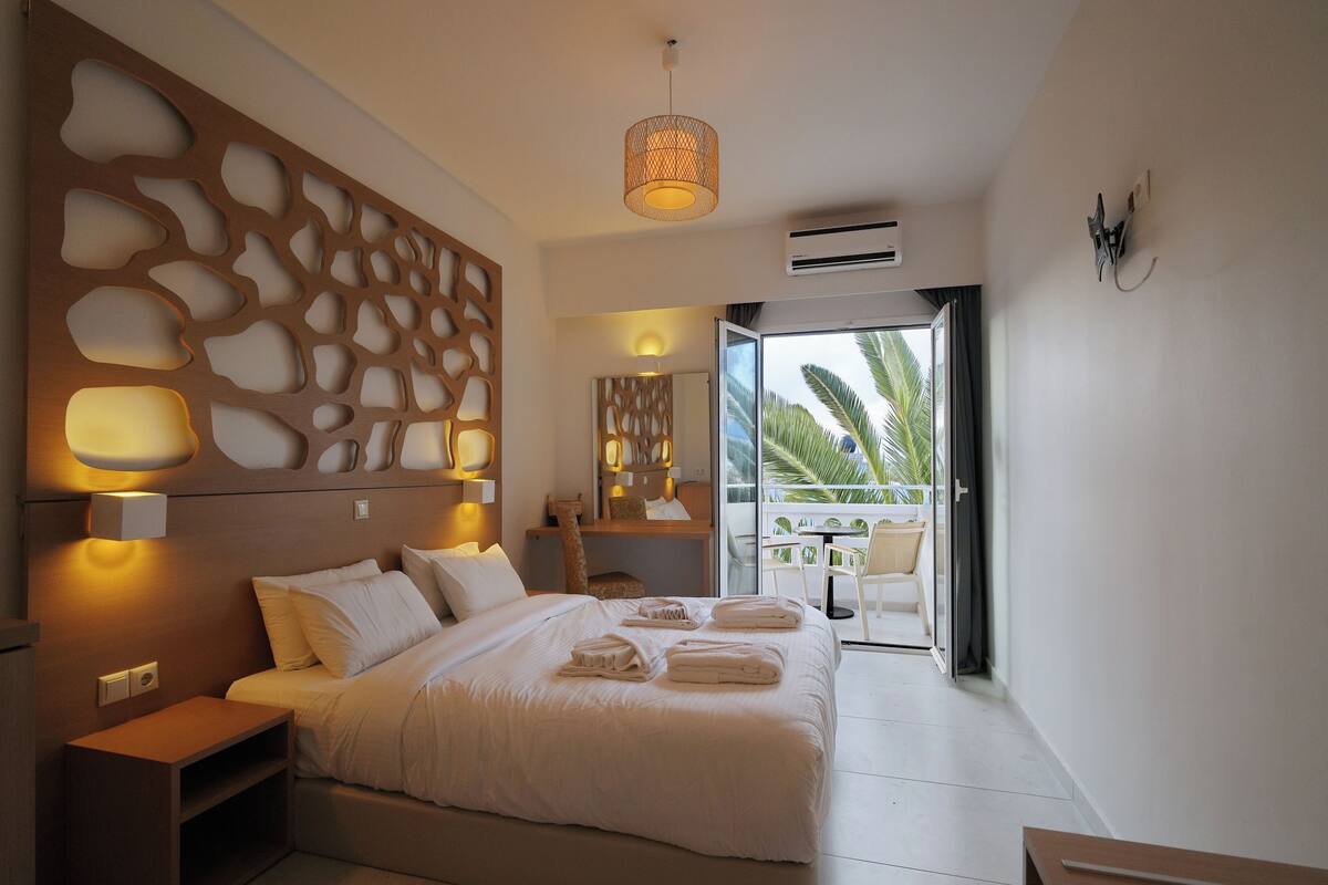 A modern hotel room with a large bed, a wooden headboard with cut-out patterns, a balcony with a table and chairs, and a view of palm trees.