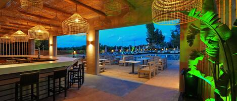 An outdoor bar area with wicker hanging lights, wooden tables and chairs, and a view of the pool in the background.