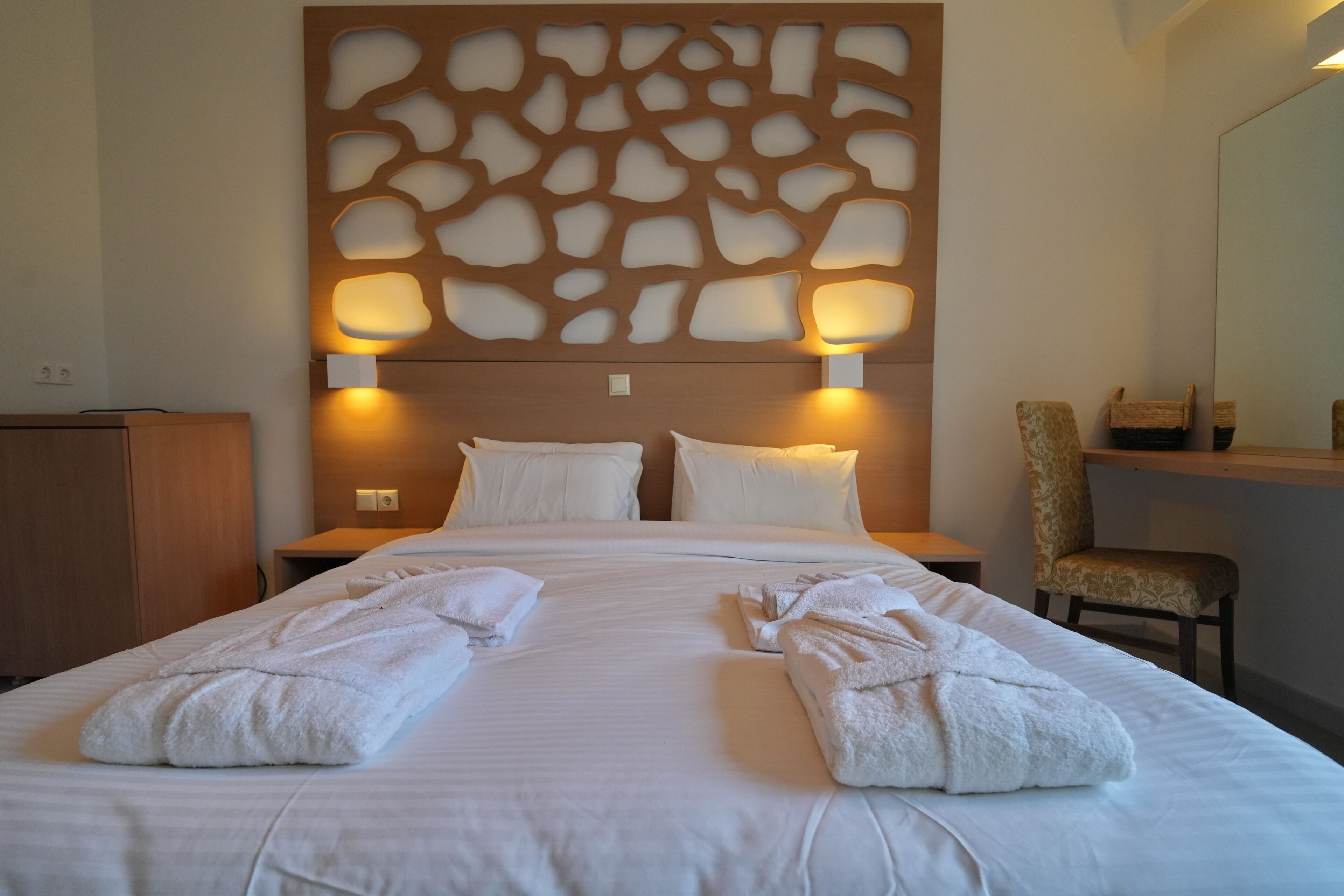 A neatly made bed with two folded towels, a wooden headboard with cut-out patterns, two bedside lamps, a wooden nightstand, and a chair with a woven seat.