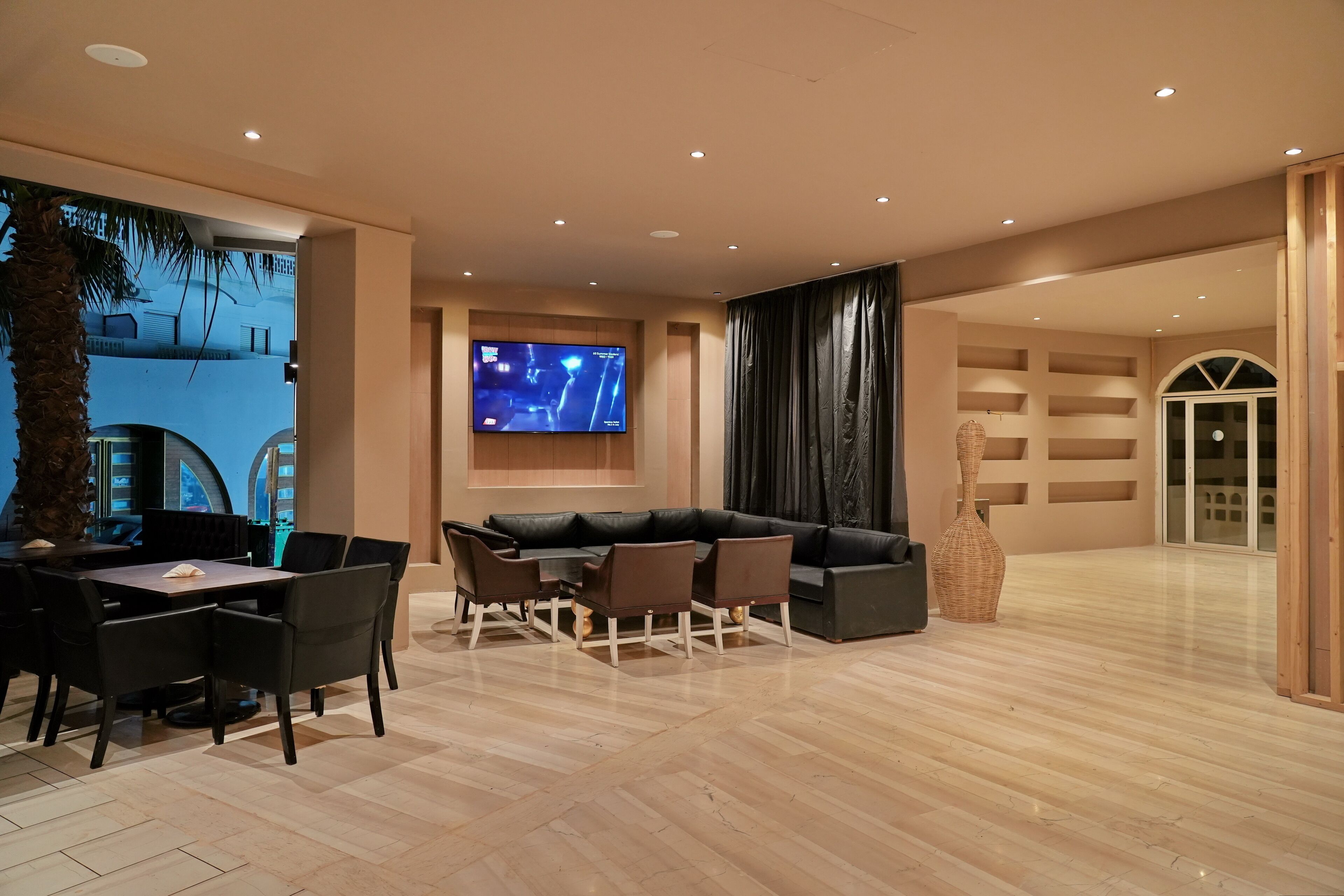 A modern living room with a large flat-screen TV mounted on the wall, a black leather sofa, and a dining area with a table and chairs.