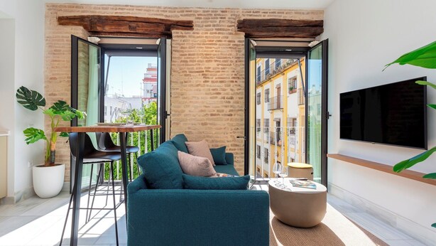 A modern living room with a sofa, a coffee table, and a TV. There is a balcony with a view of buildings and a plant. - Genteel Home San Vicente (Seville)