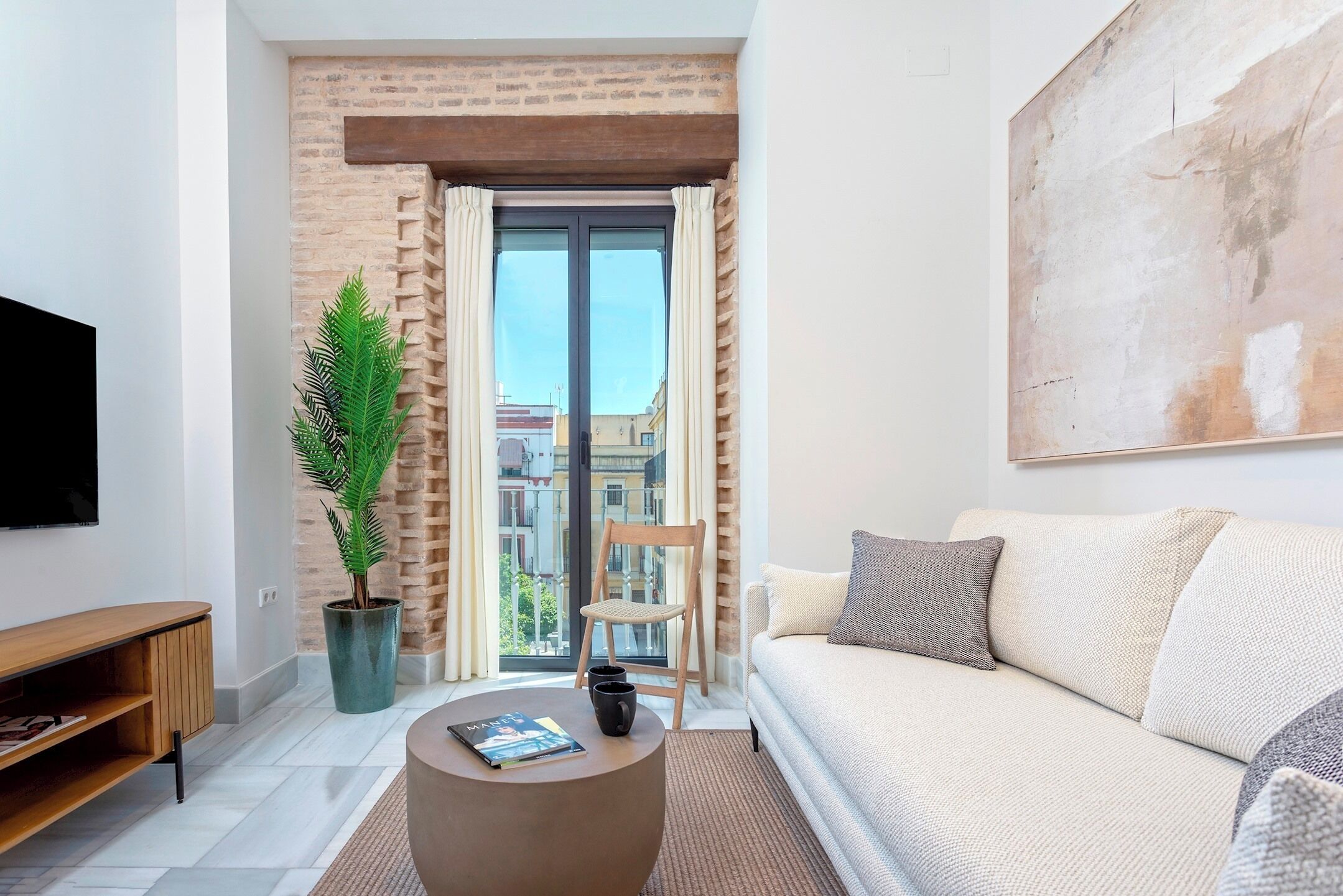 A modern living room with a sofa, a coffee table, a potted plant, and a view of the city through a large window.
