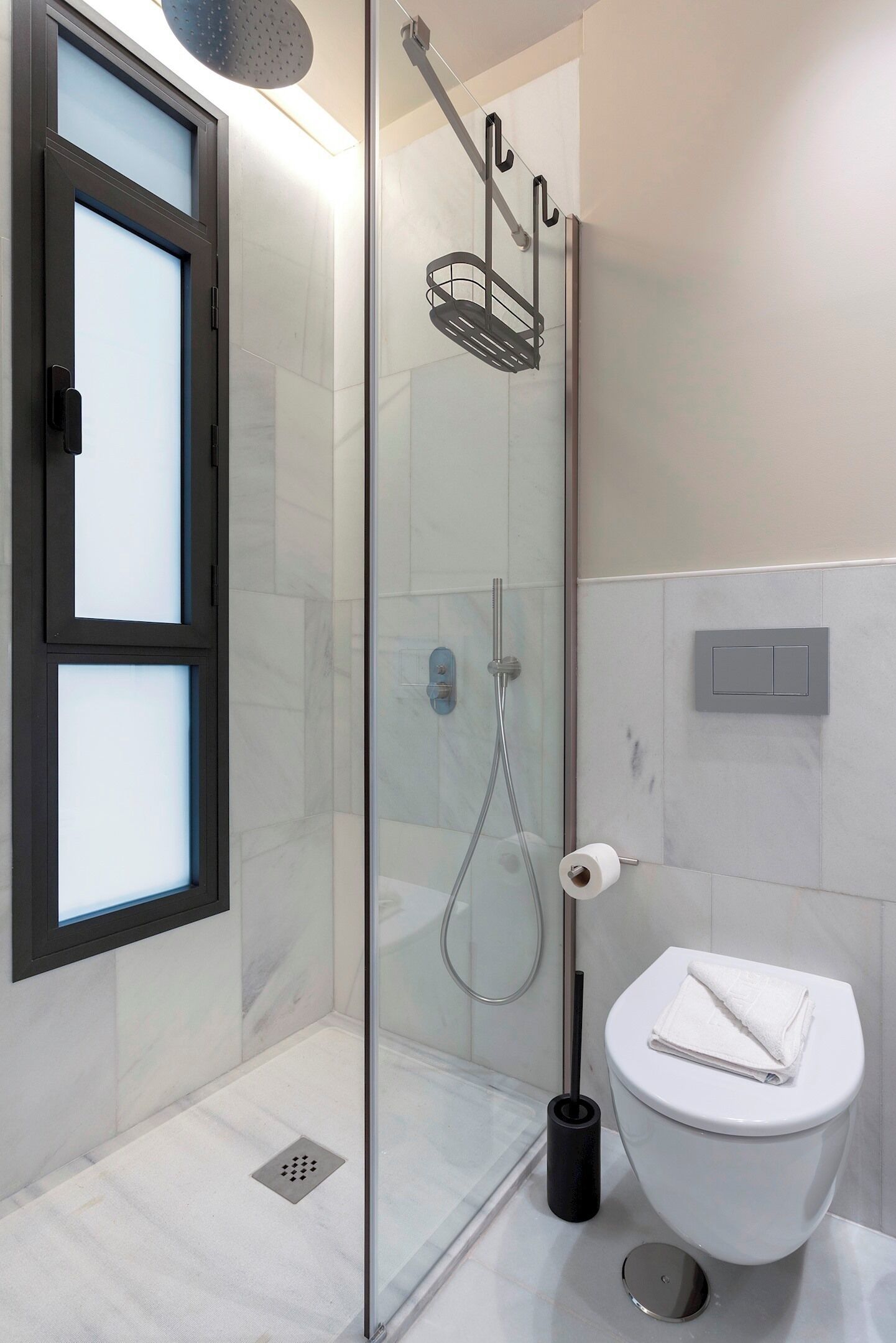 Et moderne badeværelse med en glasvæg omkring brusenichen, et hvidt toilet og et sort gulvafløb.