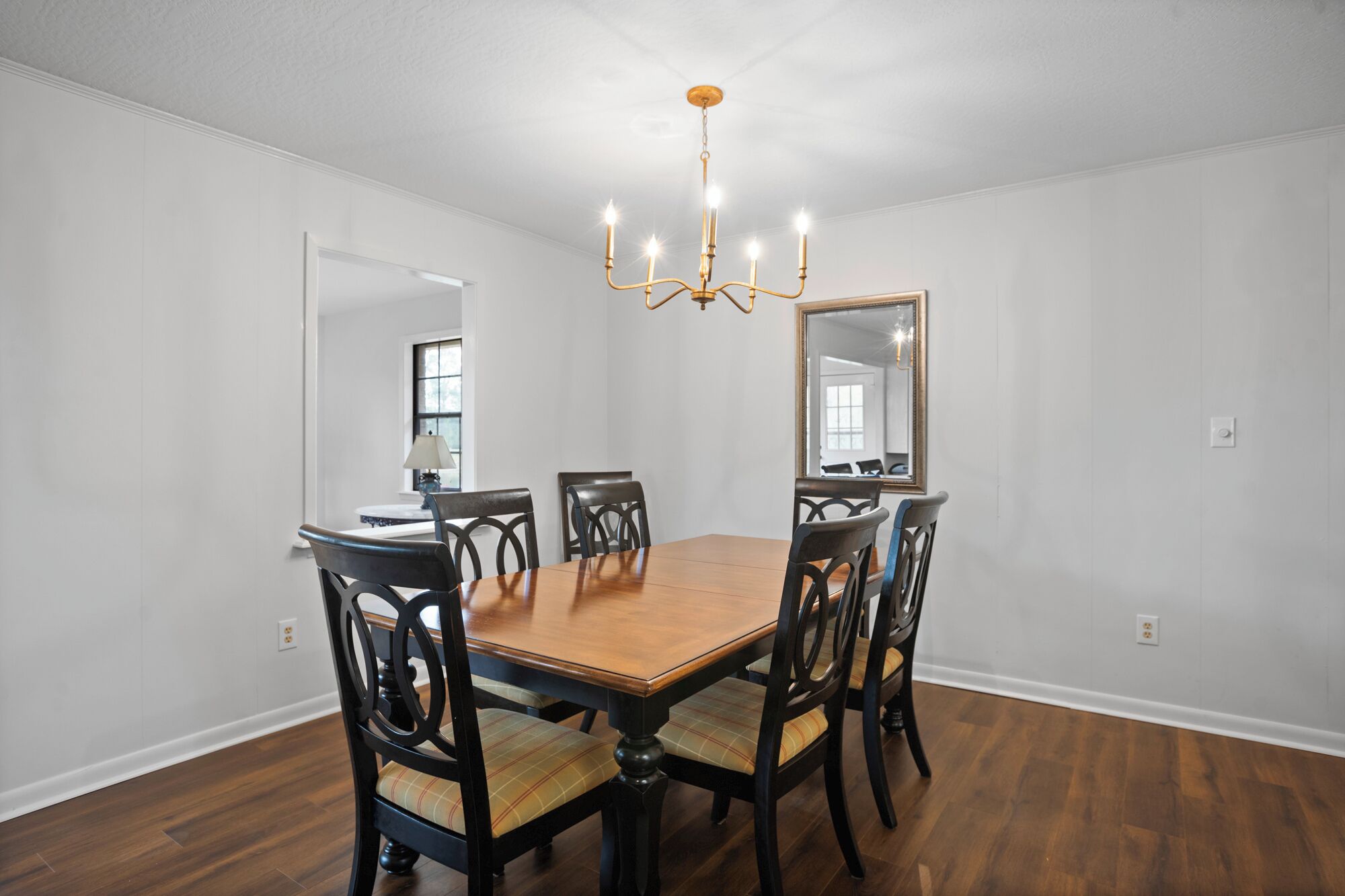 Une salle à manger avec une table et des chaises en bois, un lustre et un grand miroir au mur.