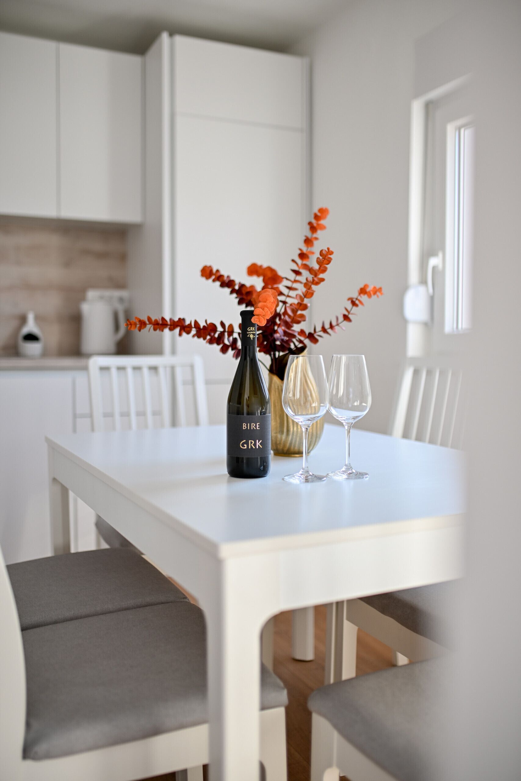 A white dining table with a bottle of wine and two glasses, accompanied by a vase of red flowers.