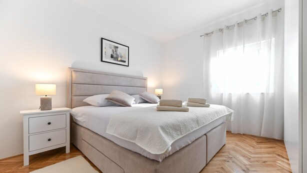 Ein modernes Schlafzimmer mit einem großen Bett, Nachttischlampen, einem gerahmten Bild an der Wand und einem Fenster mit Vorhängen.