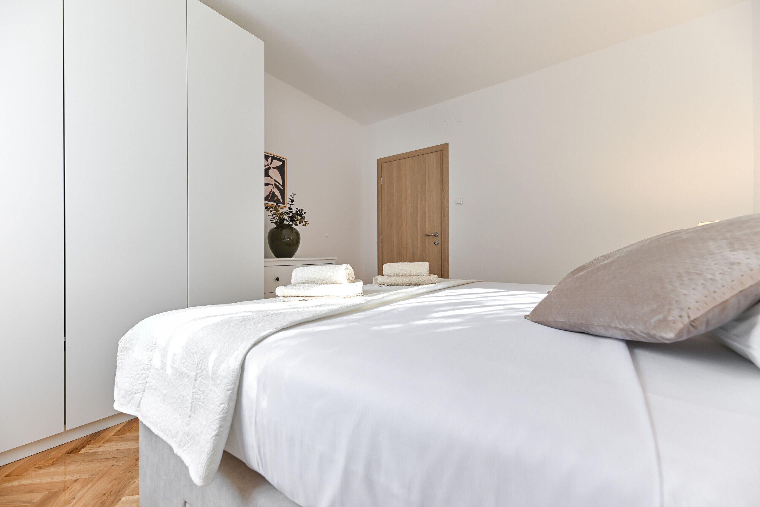 A bedroom with a bed, a wooden door, a white bedside table with a vase and towels, and a herringbone floor.