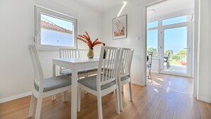 Ein Essbereich mit weißem Tisch und Stühlen, einer Vase mit orangefarbenen Blumen und einem Fenster mit Blick auf ein Dach.