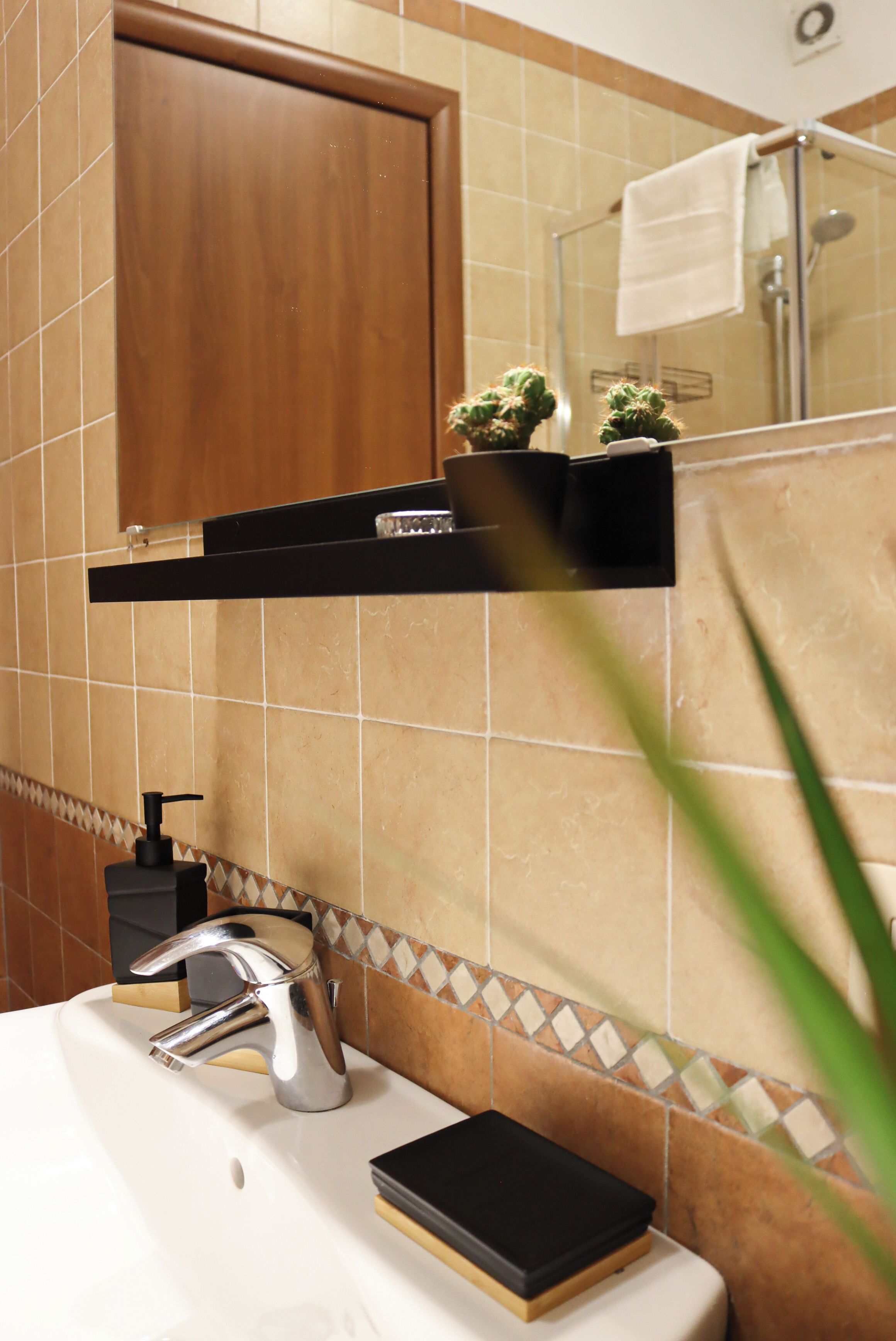 Un bagno con un lavandino bianco, un rubinetto cromato e un dispenser per sapone nero. Sopra il lavandino è presente un armadietto in legno e uno specchio con un asciugamano appeso alla porta.