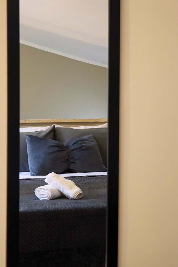 Un lit bien fait, avec une literie sombre et une serviette blanche roulée, une tête de lit et un mur sobre en arrière-plan.