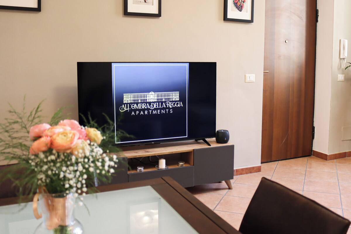 Een woonkamer met een flatscreen televisie, lijstwerken aan de muur, een houten deur en een vaas met bloemen op een tafel.