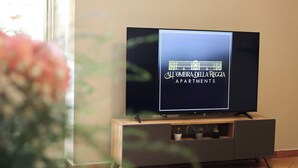 A flat-screen TV on a wooden and metal TV stand with a logo for "All'Ombra delle La Reggia Apartments". - All'Ombra Della Reggia (Caserta)