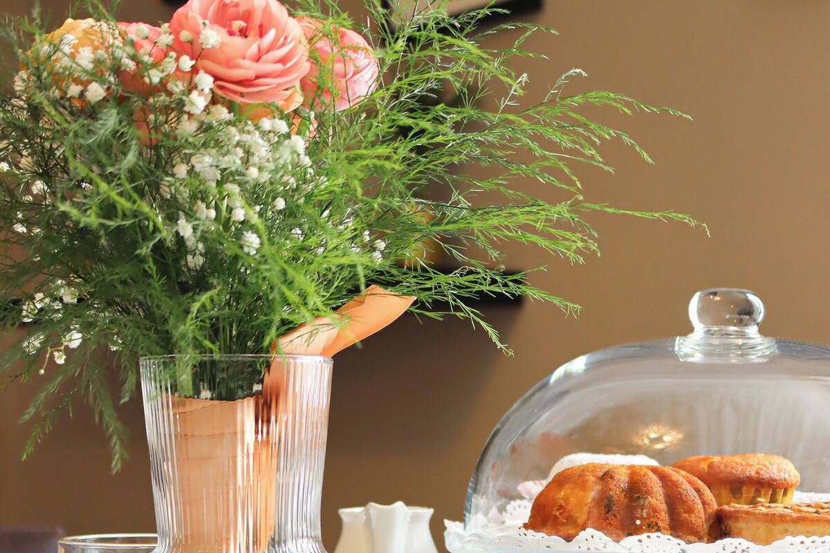 Een tafel met een glazen vaas met bloemen, een glas sinaasappelsap en een overdekte etagère met gebak.