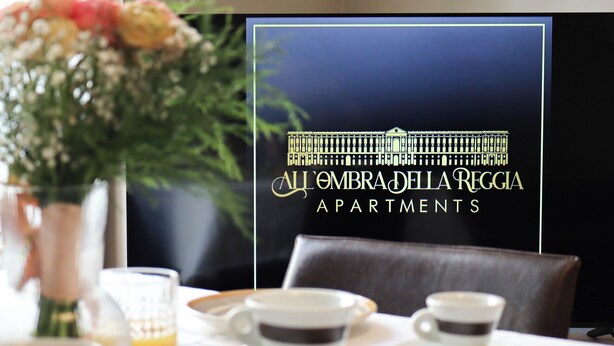A dining table with two cups of coffee and a glass of orange juice, a vase with flowers, and a framed sign for All'Ombra della Reggia Apartments. - All'Ombra Della Reggia (Caserta)