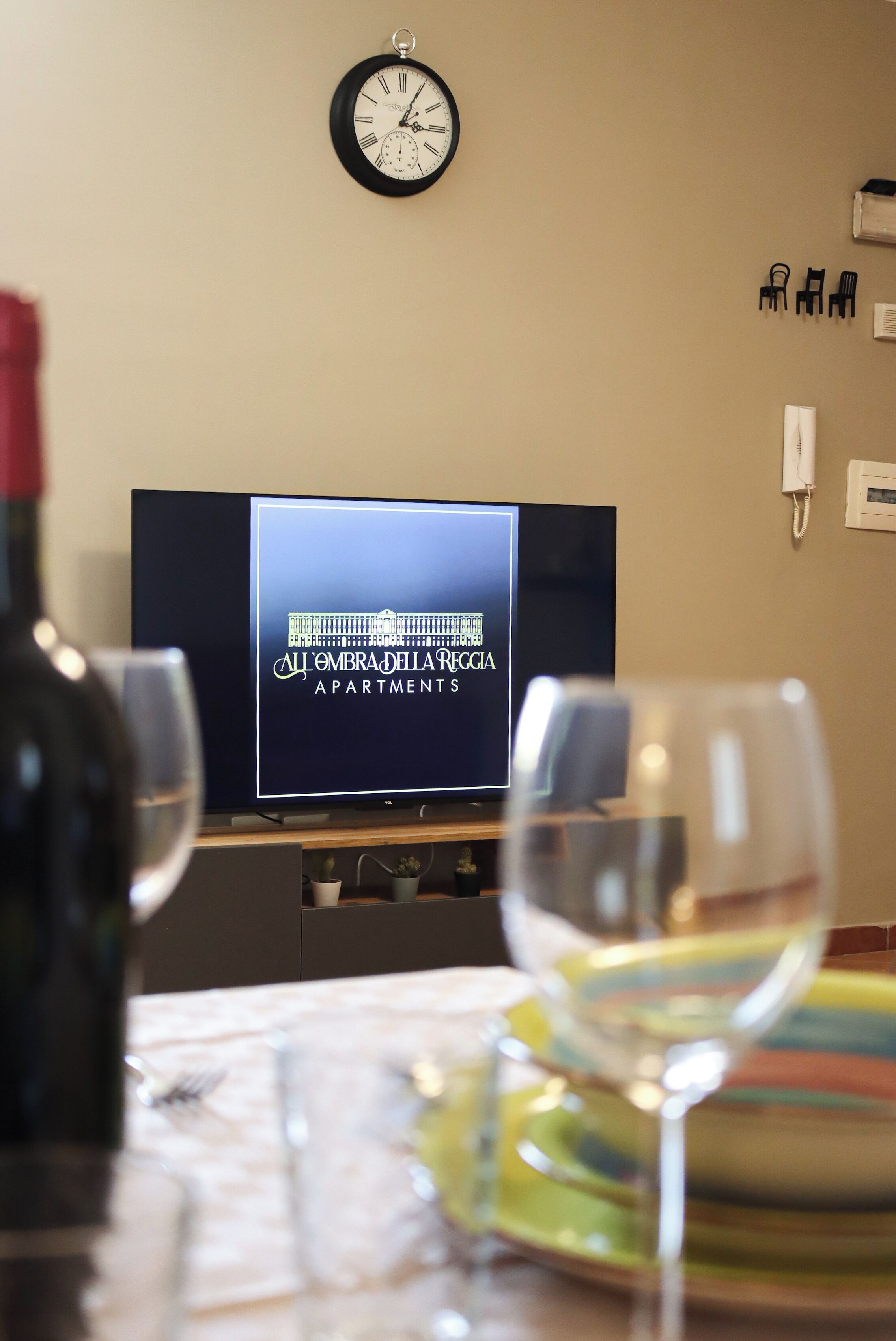 A room with a television displaying 'All Embra' apartments, a clock on the wall, and glasses on a table.