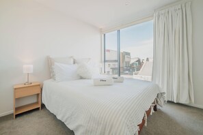 Habitación de hotel con una cama grande, una mesita de noche, una lámpara y vistas a la ciudad a través de un ventanal amplio.