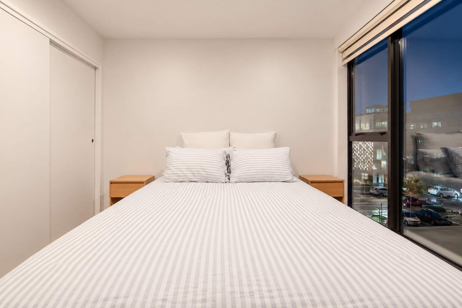 A bedroom with a bed, two nightstands, and a large window with a city view.
