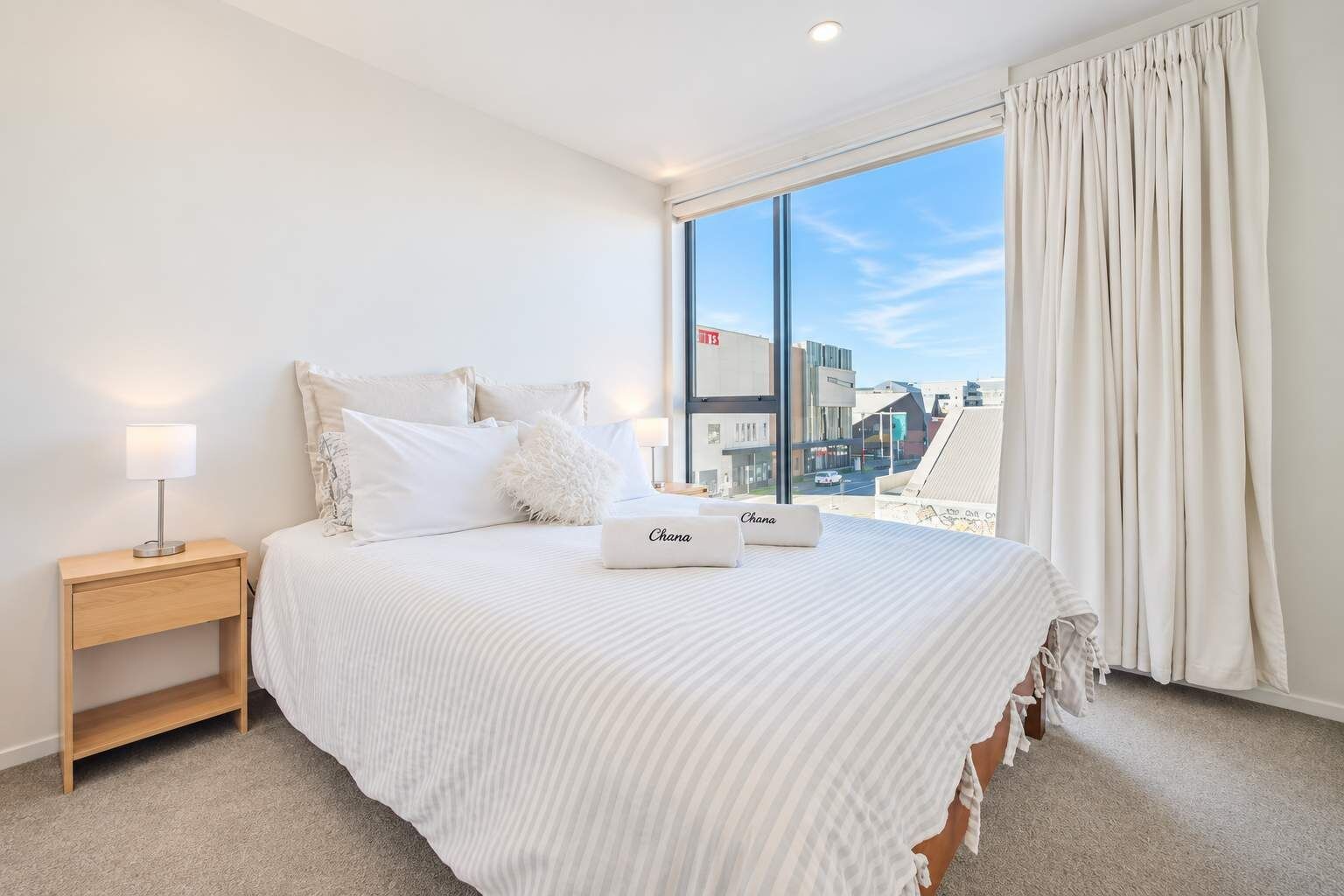 A hotel room with a large bed, a nightstand, a lamp, and a view of the city through a window.