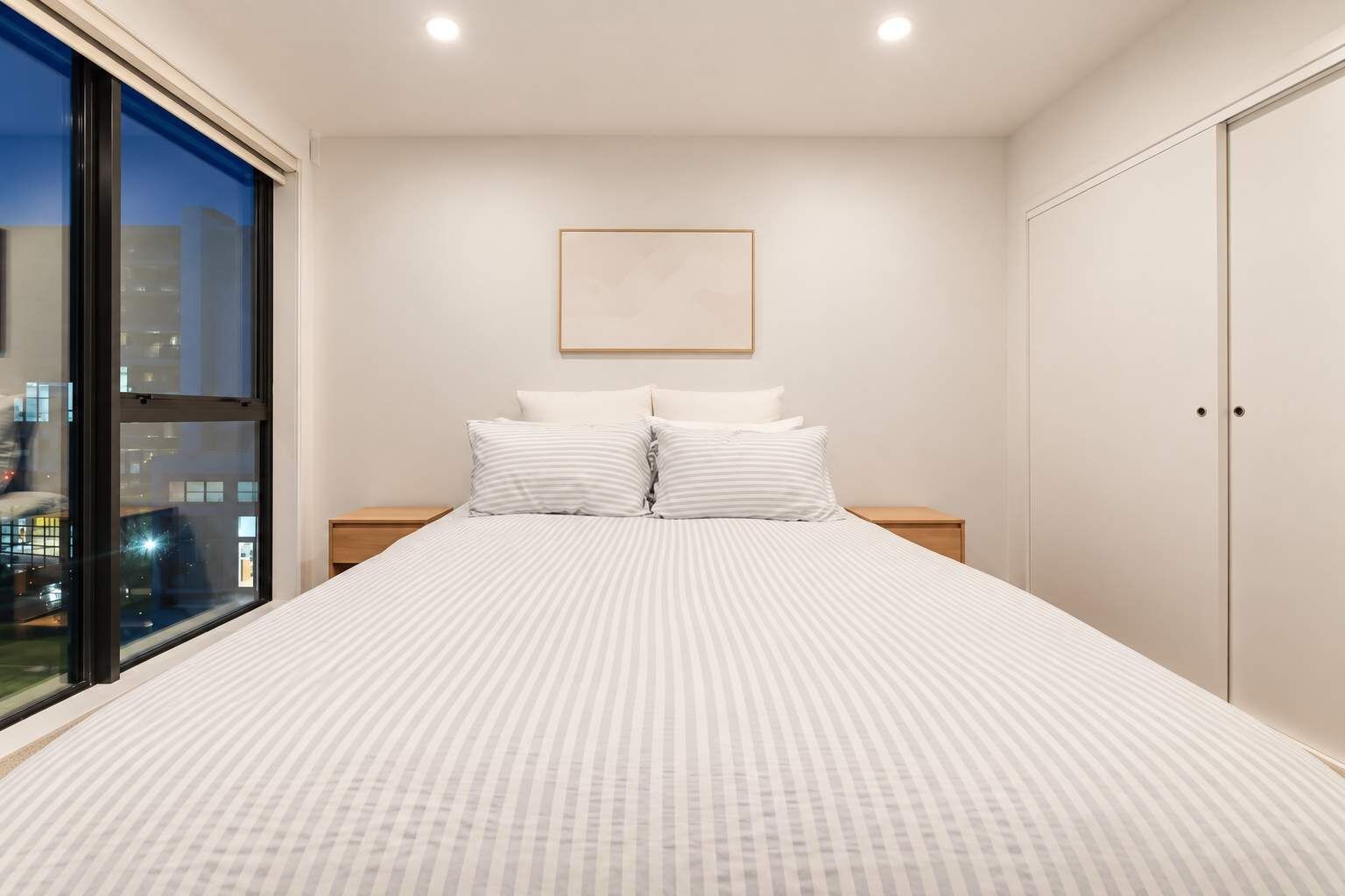 A bedroom with a large bed, two nightstands, a window with a city view, and a wardrobe.