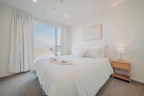 A hotel room with a large bed, white bedding, a nightstand with a lamp, and a view of the sea through a large window.