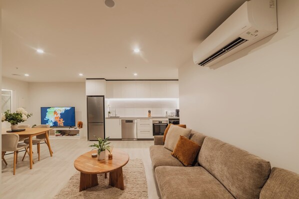 Una sala moderna con sofá, mesa de centro y televisor. Se ve una cocina con armarios y electrodomésticos blancos al fondo.