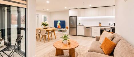 Una sala moderna con un sofá, una mesa de centro y un televisor. Hay una zona de cocina con armarios blancos y un refrigerador.