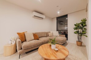 A modern living room with a beige sectional sofa, a wooden coffee table, a side table with a vase and a plant, and a large window with a view of the kitchen.